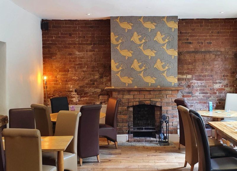 Interior shot of The Fish, Wixford. Showing a cosy atmosphere, with several tables surrounded by comfy chairs, and a fireplace embedded in a brick work wall.